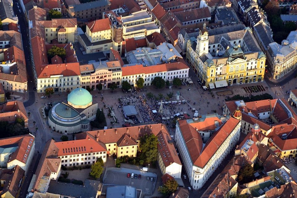 Una vista aerea di una piazza europea affollata, caratterizzata da una grande cupola verde e edifici con tetti tradizionali in tegole rosse.