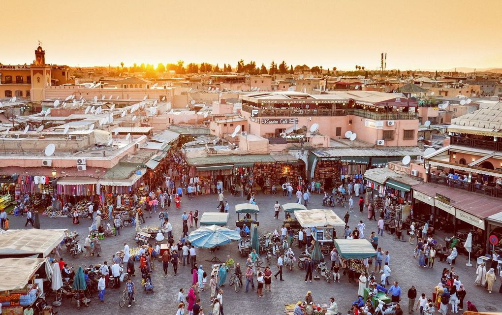An elevated view of a bustling city marketplace with many people, stalls, and terracotta buildings at sunset.