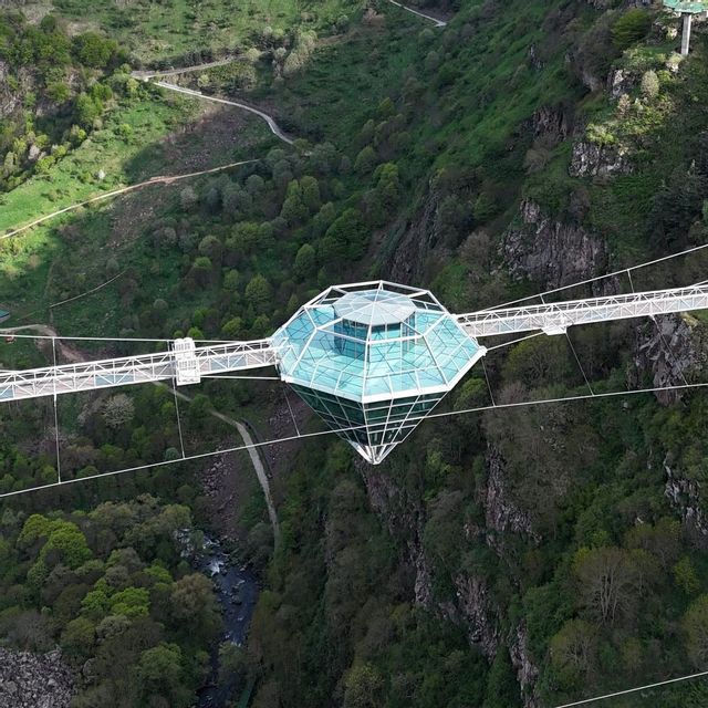 Vue aérienne d'un pont suspendu blanc avec un point de vue en verre en forme de diamant au-dessus d'un canyon profond et boisé.