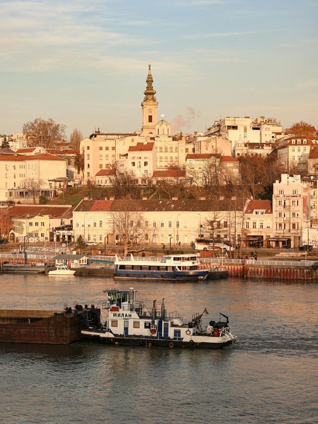 Un rimorchiatore manovra una chiatta su un fiume, con una città collinare e un campanile di chiesa prominente sullo sfondo.