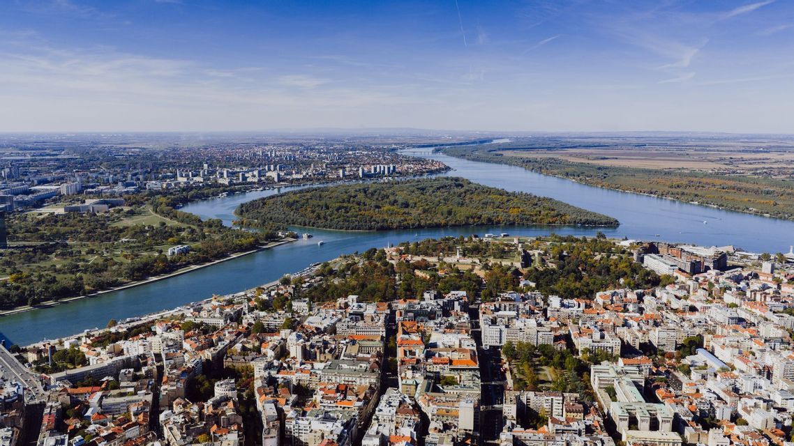 Vista aerea di una vasta città con un ampio fiume che si biforca, scorrendo attorno a una grande isola boscosa sotto un cielo azzurro.
