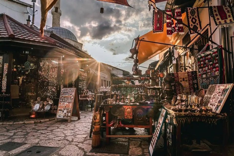Bancarelle di artigianato tradizionale, oggetti in metallo e tessuti fiancheggiano una strada acciottolata illuminata dal sole in un bazar, con la cupola di una moschea sullo sfondo.