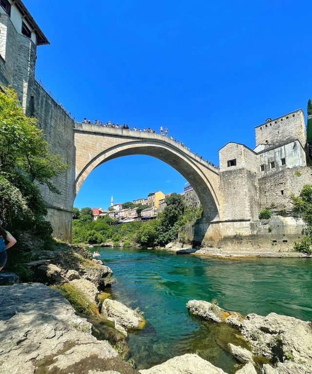 Prospettiva dal basso di un grande ponte ad arco in pietra che attraversa un fiume turchese, con persone riunite in cima sotto un cielo azzurro e limpido.