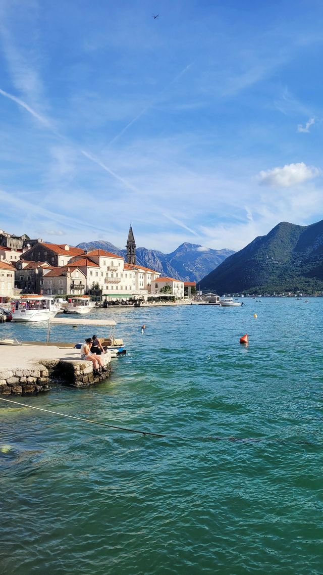 Due persone sedute su un molo di pietra che si affaccia su una baia con una città costiera e montagne sullo sfondo.