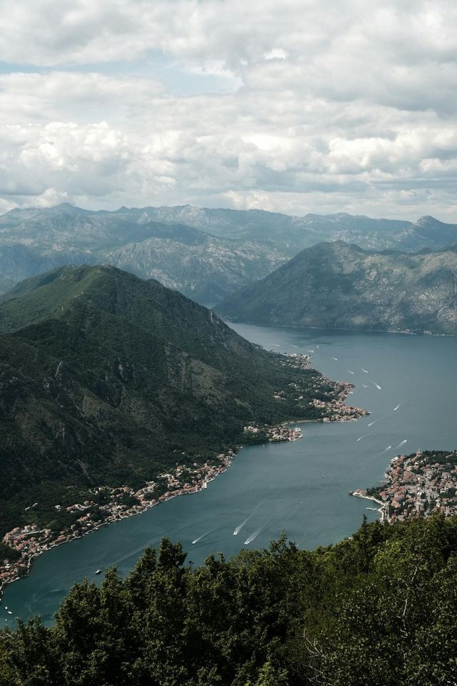 Una vista aerea di una baia sinuosa circondata da montagne verdi, con paesini lungo la riva e barche sull'acqua.