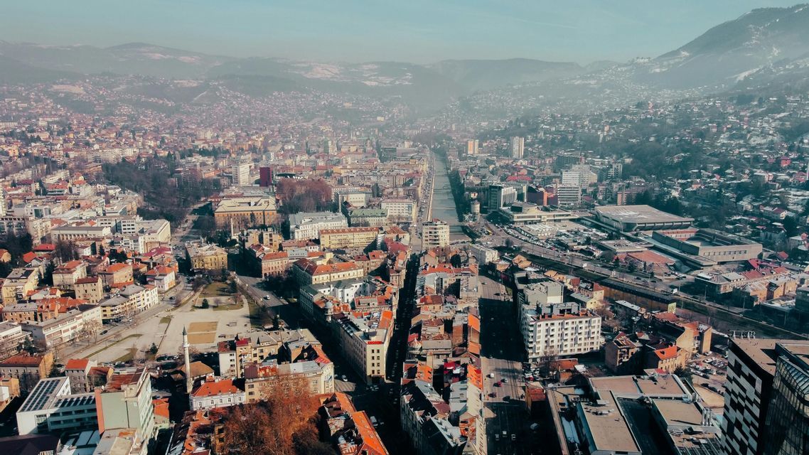 Una veduta aerea di una città fitta con edifici dai tetti rossi e un fiume, che si estende verso montagne sfumate all'orizzonte.