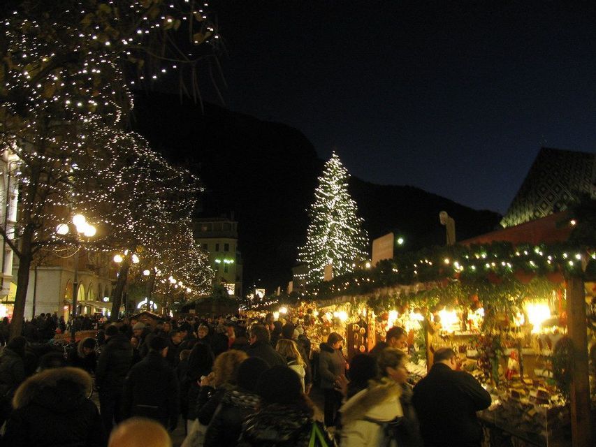 Una grande folla di persone cammina attraverso un mercatino di Natale all'aperto di notte, con bancarelle illuminate e alberi decorati.