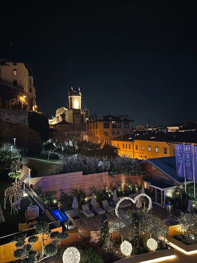 Una terrazza panoramica con lettini, luci decorative e statue di giraffa, con vista su un castello illuminato e sul paesaggio urbano notturno.