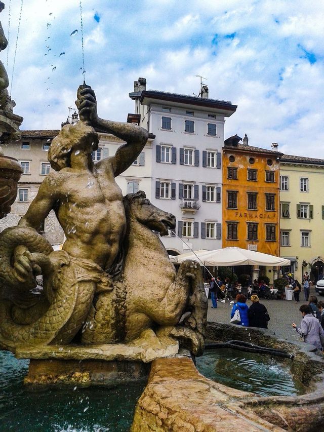 Una fontana in pietra con una statua di un tritone a cavallo di un cavalluccio marino in una piazza di città europea con edifici colorati.