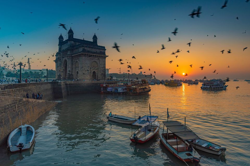 Pájaros sobrevuelan barcos en el agua, frente a un gran monumento de arco de piedra, durante un atardecer colorido.