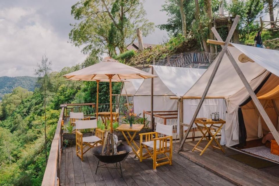 Un campamento de glamping en una terraza de madera con tiendas blancas, sillas, una sombrilla y una hoguera con vistas a un bosque exuberante.