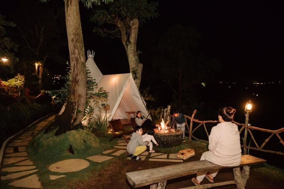 Un grupo de personas se reúne alrededor de una fogata por la noche junto a una tienda tipi iluminada en una zona boscosa.