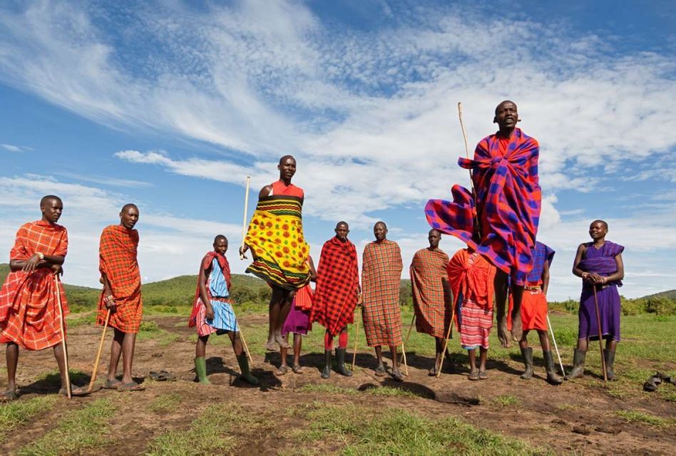 Un gruppo di Maasai in abiti tradizionali colorati in un campo, con due uomini che saltano in alto.