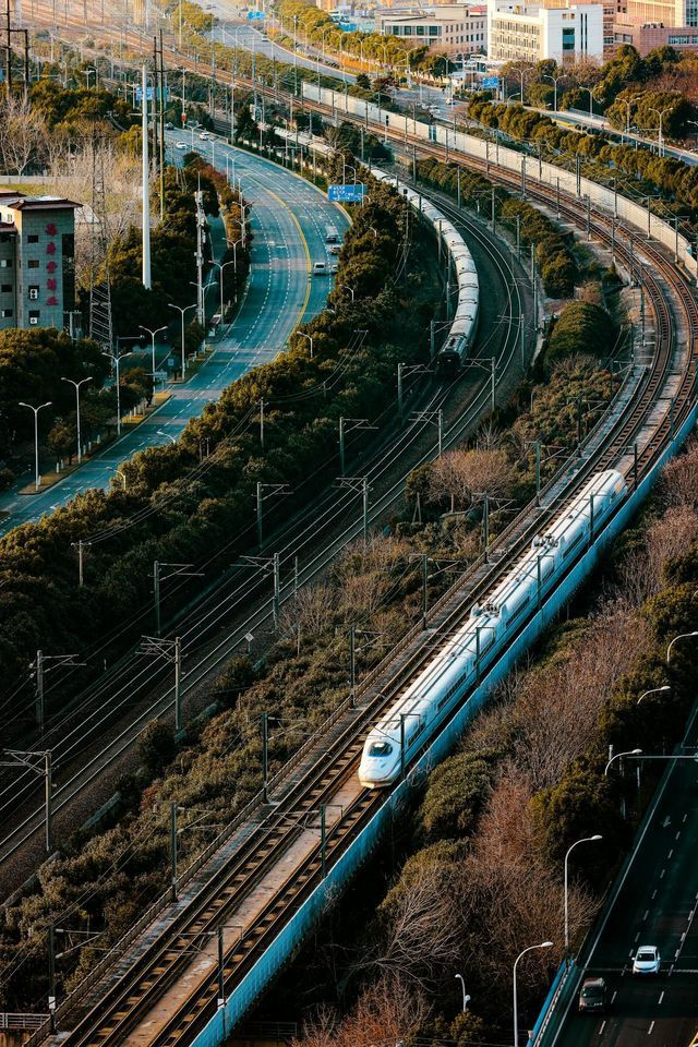 Veduta aerea di un treno proiettile bianco che viaggia su binari curvi, affiancato da un'autostrada tortuosa e altre linee ferroviarie.