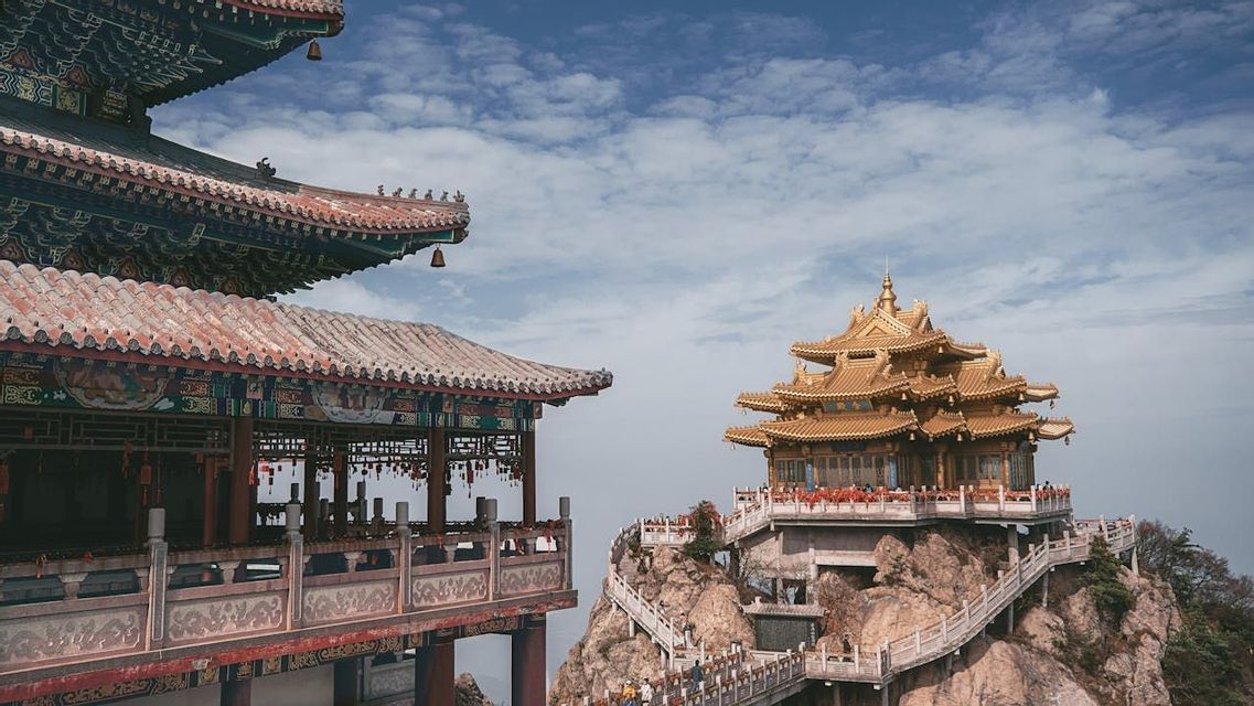 Un tempio dorato sorge su una montagna rocciosa, collegato da un ponte a un altro edificio tradizionale sotto un cielo azzurro e nuvoloso.