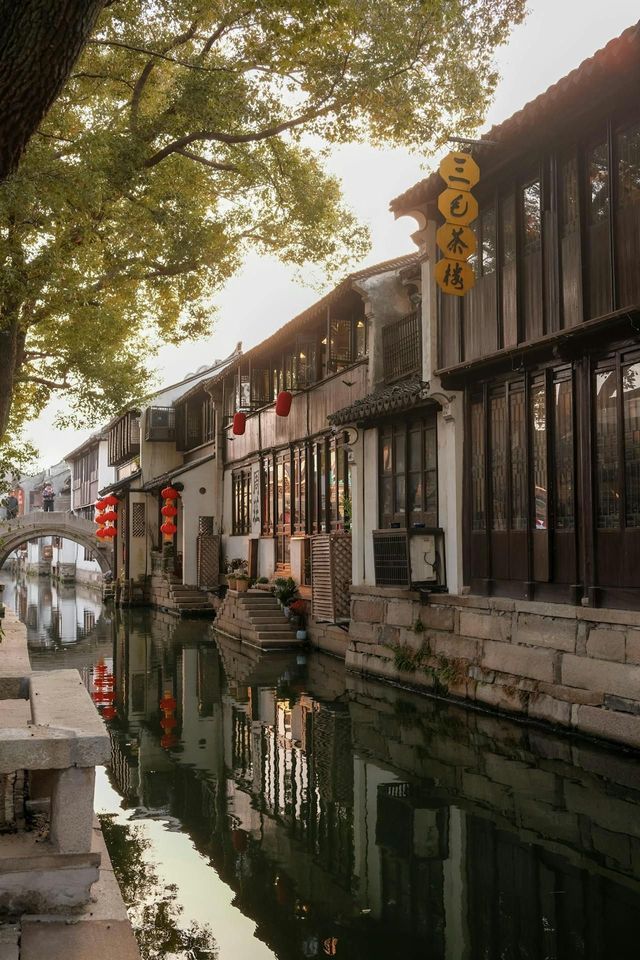 Edifici tradizionali con lanterne rosse costeggiano un canale calmo, con i loro riflessi che luccicano nell'acqua sotto un ponte in pietra ad arco.
