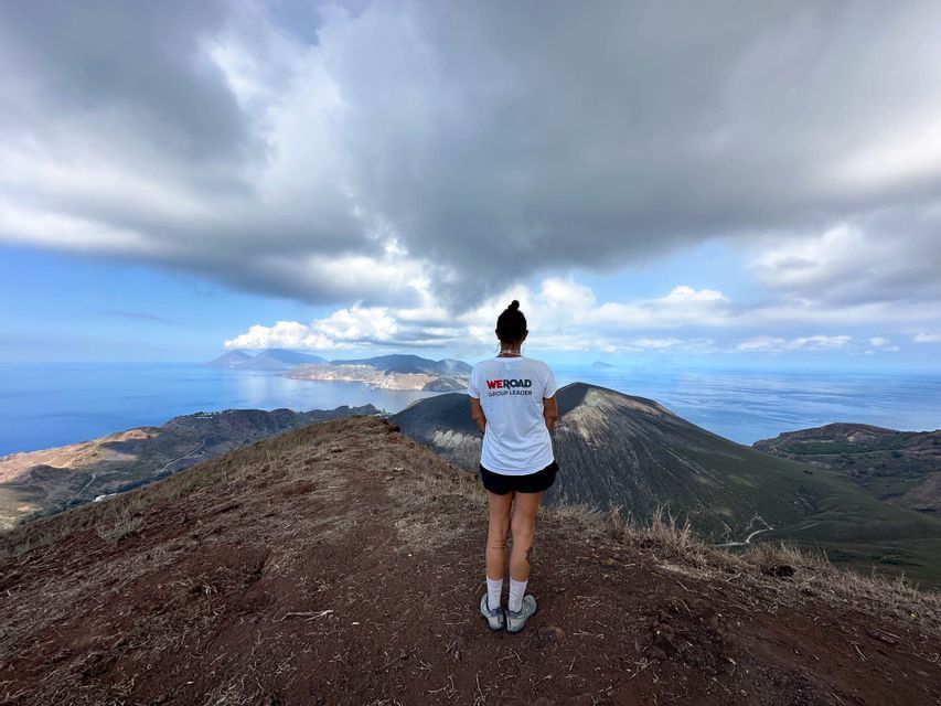 Un capogruppo WeRoad si trova su una cresta di montagna che domina un paesaggio vulcanico e il mare sotto un cielo nuvoloso.