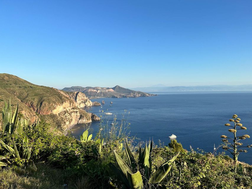 Blick von einem grünen Hügel auf eine felsige Küste, ein tiefblaues Meer mit Booten und Vulkaninseln am Horizont.