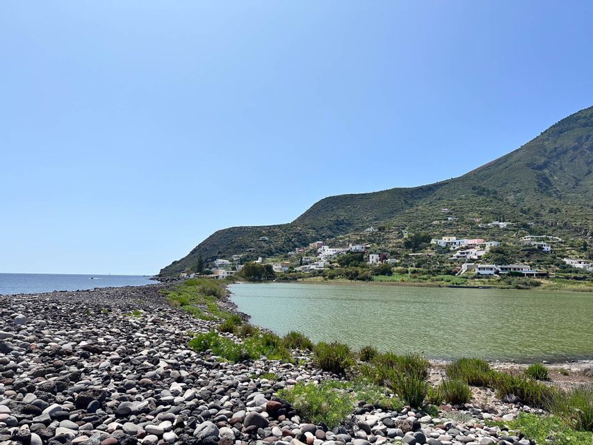 Ein Kieselstrand trennt das blaue Meer von einer grünen Lagune, mit einem Dorf auf einem üppig grünen Hügel unter klarem Himmel.