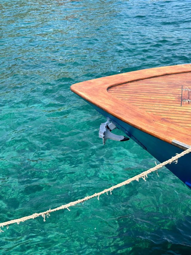 La prua di una barca con coperta in legno galleggia su acqua turchese trasparente, con l'ancora sollevata.