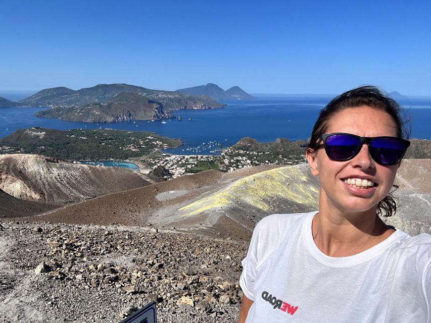 Una donna sorridente con una maglietta WeRoad e occhiali da sole si scatta un selfie da una cima vulcanica che si affaccia su una baia con isole.