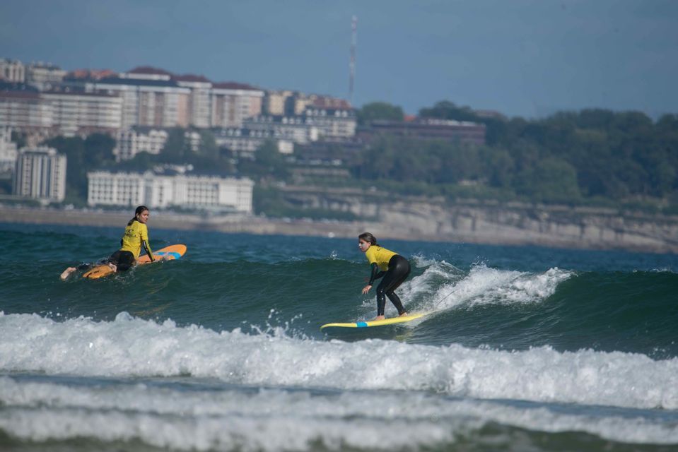 Dos personas de un viaje en grupo de WeRoad surfeando olas, con una en tabla de surf y una costa urbana de fondo.