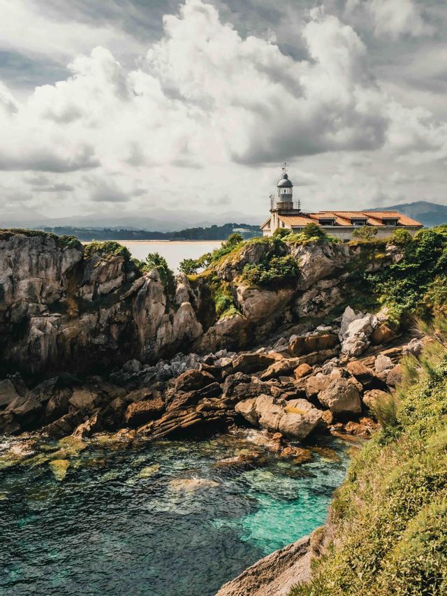 Un faro con techo de tejas se asienta en un acantilado rocoso y verde, con vistas a aguas claras y turquesas bajo un cielo nublado.