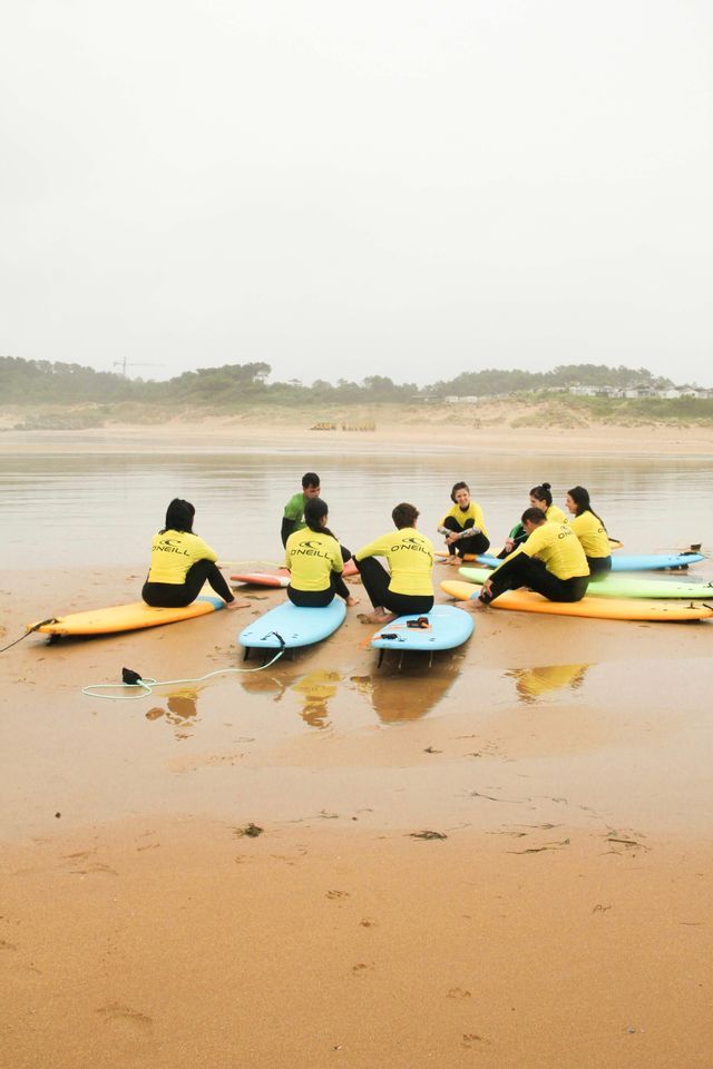 Un grupo de WeRoad con camisetas amarillas se sienta en tablas de surf en círculo en una playa para una clase de surf.