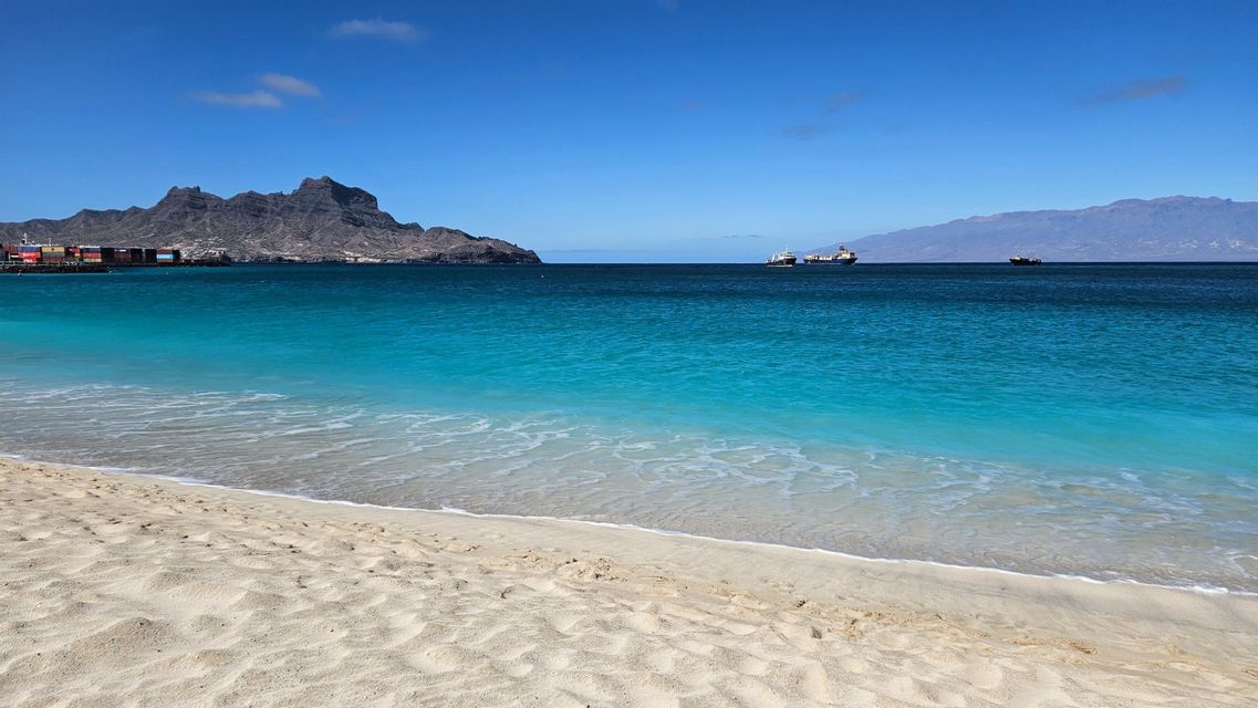 Ein Sandstrand mit leuchtend türkisfarbenem Wasser trifft auf ein tiefblaues Meer, mit felsigen Bergen und Schiffen in der Ferne.