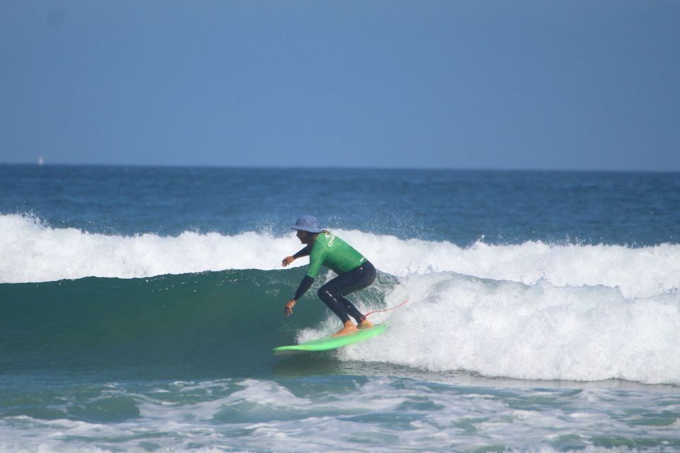 Un surfista con un traje de neopreno verde y negro y gorra azul surfea una ola en una tabla de surf verde brillante en el océano.