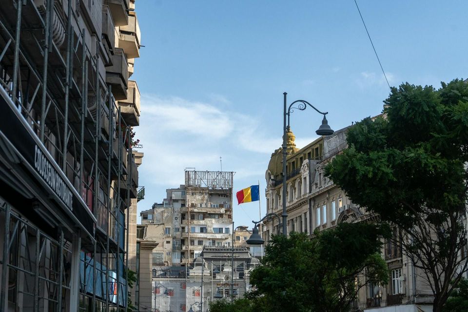 Eine rumänische Flagge hängt an einer Stadtstraße, umrahmt von alter, verzierter Architektur und einem weiteren Gebäude mit Gerüst.