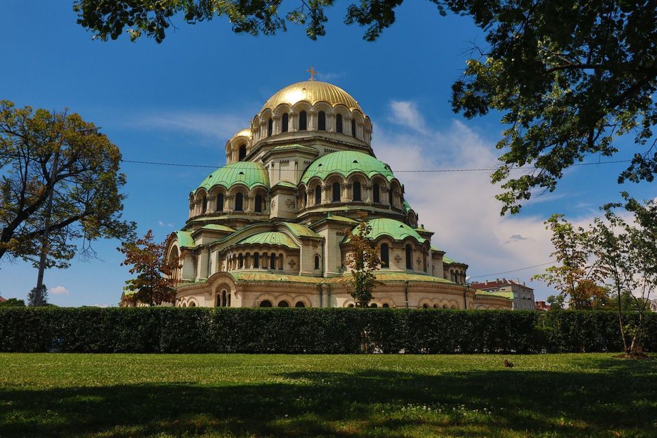 Eine große Kathedrale mit goldener Kuppel und grünen Dächern, von einem Park mit Hecke unter klarem blauem Himmel gesehen.