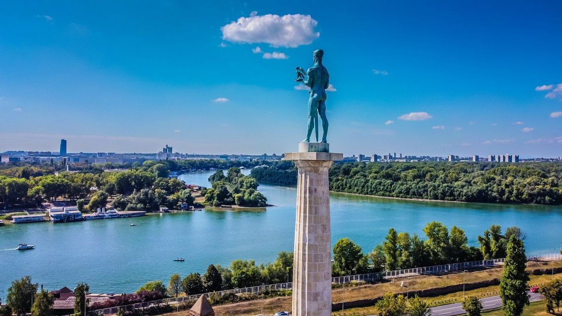 Eine Bronzestatue eines Kriegers auf einer hohen Säule blickt auf einen breiten Fluss, bewaldete Ufer und eine ferne Skyline unter blauem Himmel.