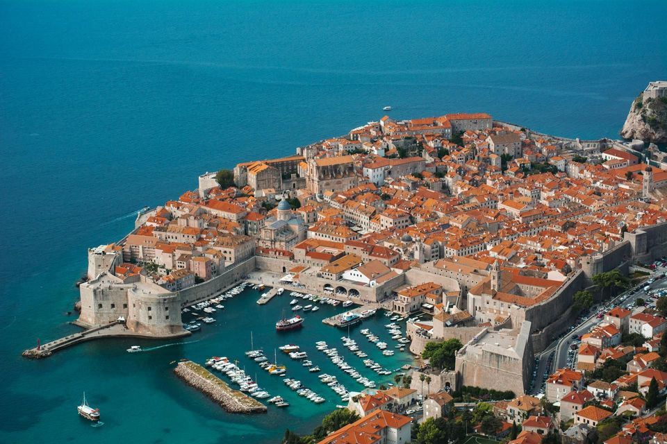 Luftaufnahme einer historischen, ummauerten Stadt mit orangefarbenen Ziegeldächern auf einer Halbinsel, die einen kleinen Hafen voller Boote im blauen Meer zeigt.