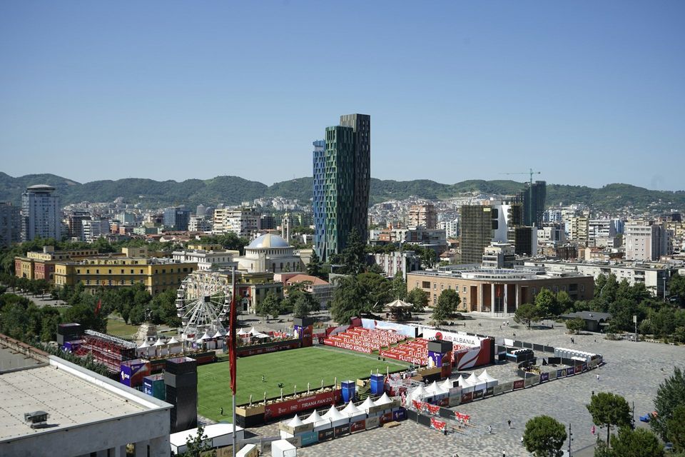 Eine Vogelperspektive einer Stadtlandschaft mit einer Fanzone auf einem Platz, die einen Fußballplatz, einen modernen Wolkenkratzer und ein Riesenrad zeigt.