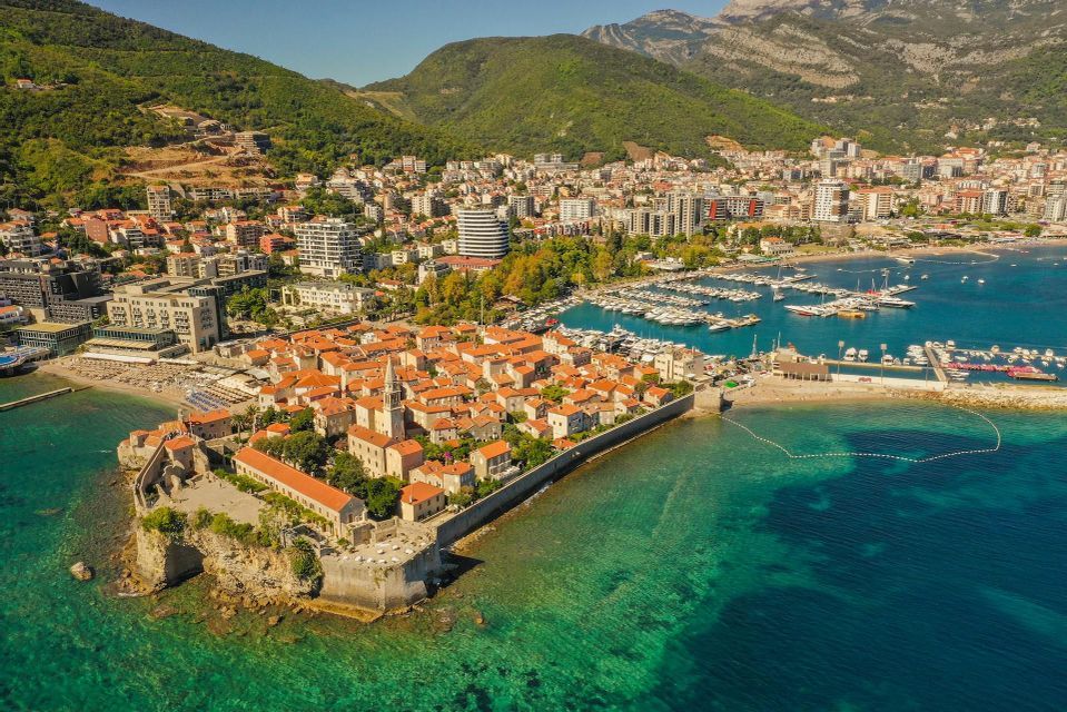 Eine Luftaufnahme einer ummauerten Küstenstadt auf einer Halbinsel, umgeben von türkisfarbenem Wasser, mit einem Yachthafen und Bergen in der Nähe.