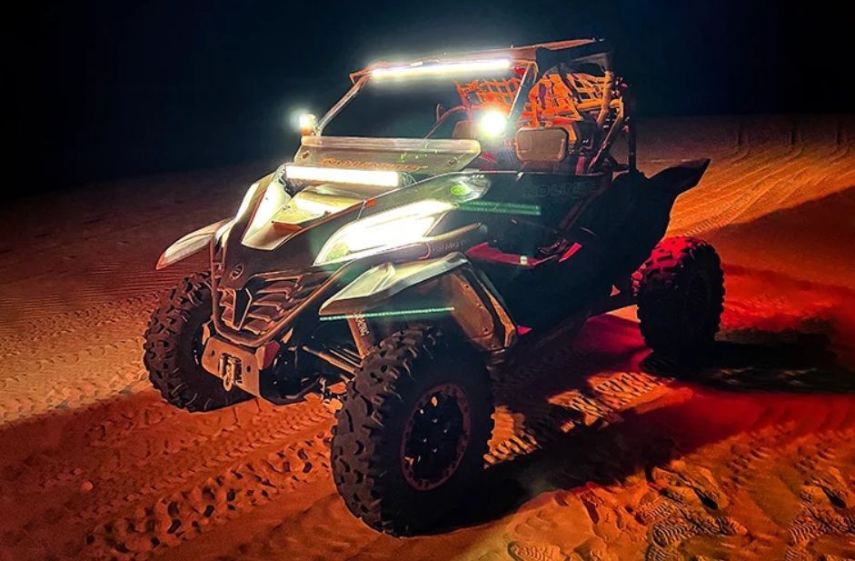 Un buggy de dunas está aparcado en la arena de noche con sus potentes faros y barras de luces encendidos, iluminando el terreno.