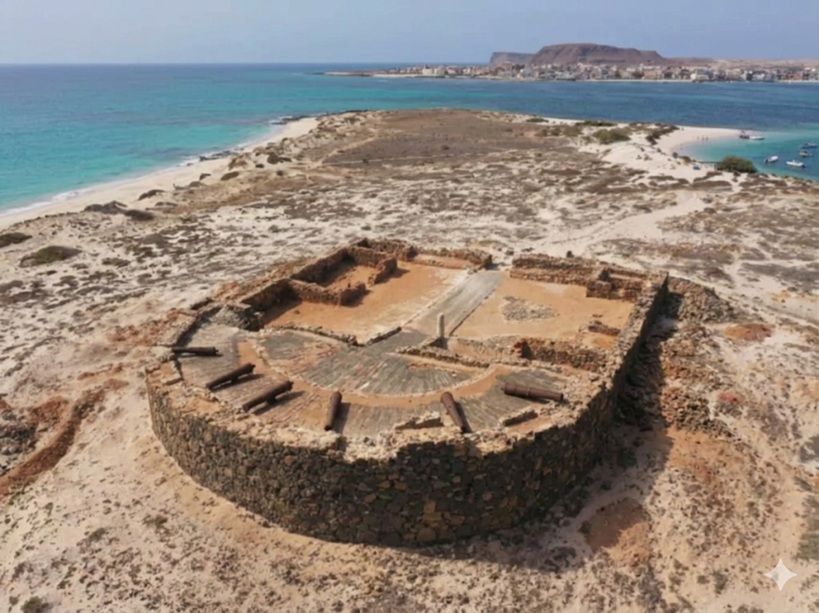 Vista aérea de las ruinas de un fuerte de piedra circular con cañones en una península arenosa, con océano turquesa y una ciudad distante.