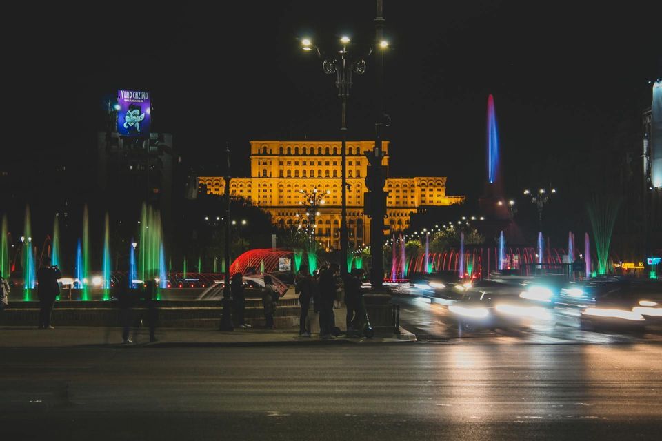 Un imponente edificio illuminato di notte, con fontane colorate e scie luminose a lunga esposizione delle auto su una strada di città.