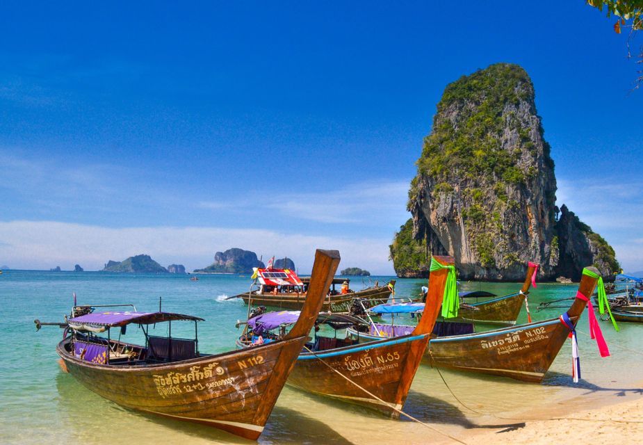 Des bateaux à longue queue en bois aux rubans colorés sont amarrés sur une plage de sable fin aux eaux turquoises, avec une île karstique calcaire en arrière-plan.