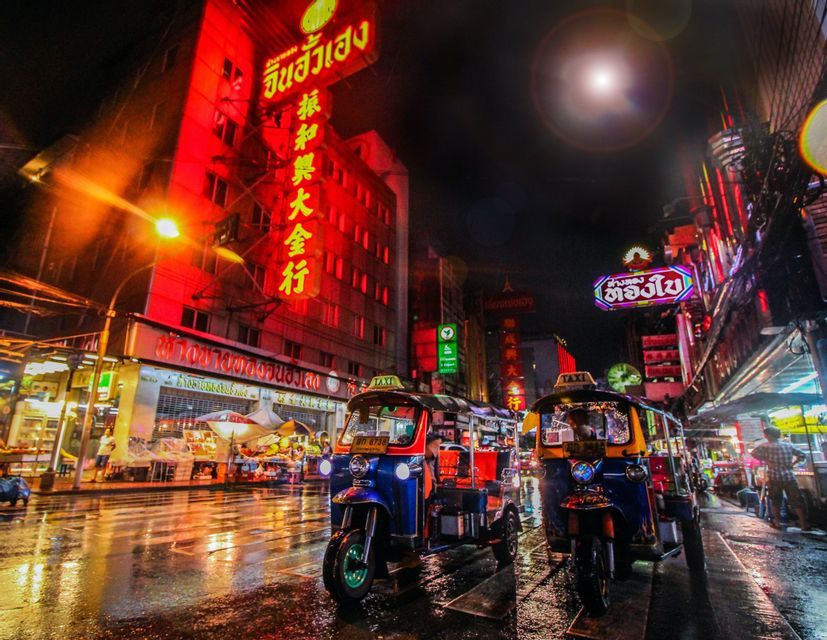 Two tuk-tuks parked on a wet city street at night, with buildings illuminated by glowing red and multicolored neon signs.