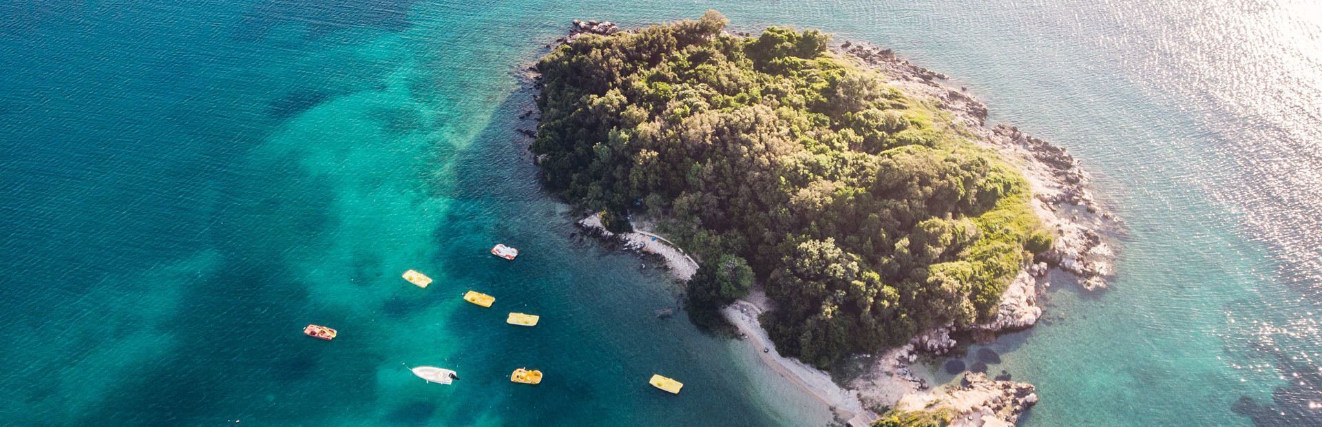 An aerial view of a small, tree-covered island in turquoise waters, with several yellow pedal boats floating nearby.