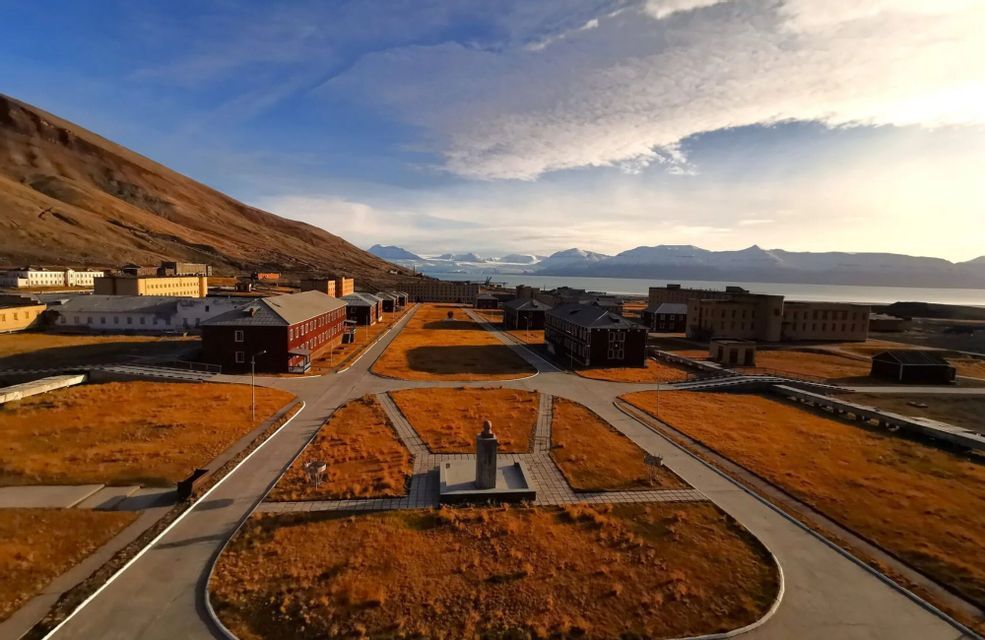 Una vista aérea de un pueblo ártico con edificios y carreteras, situado junto a una montaña, con un fiordo y glaciares al fondo.