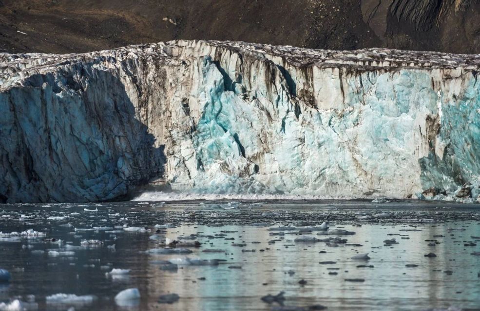 Un gran muro glaciar de hielo azul y blanco se alza sobre aguas oscuras, con pequeños icebergs flotando.