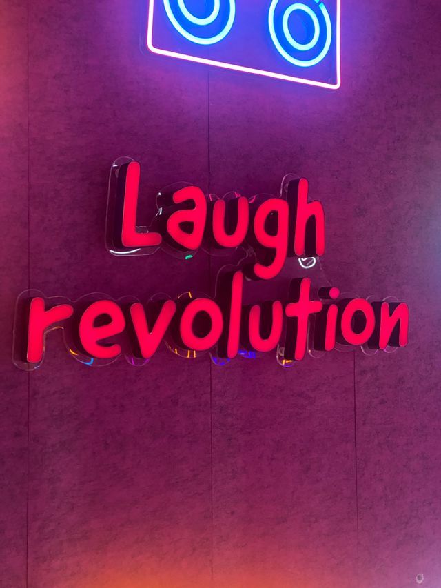Un letrero rojo estilo neón dice 'Laugh revolution' en una pared morada texturizada, con una forma de neón azul encima.