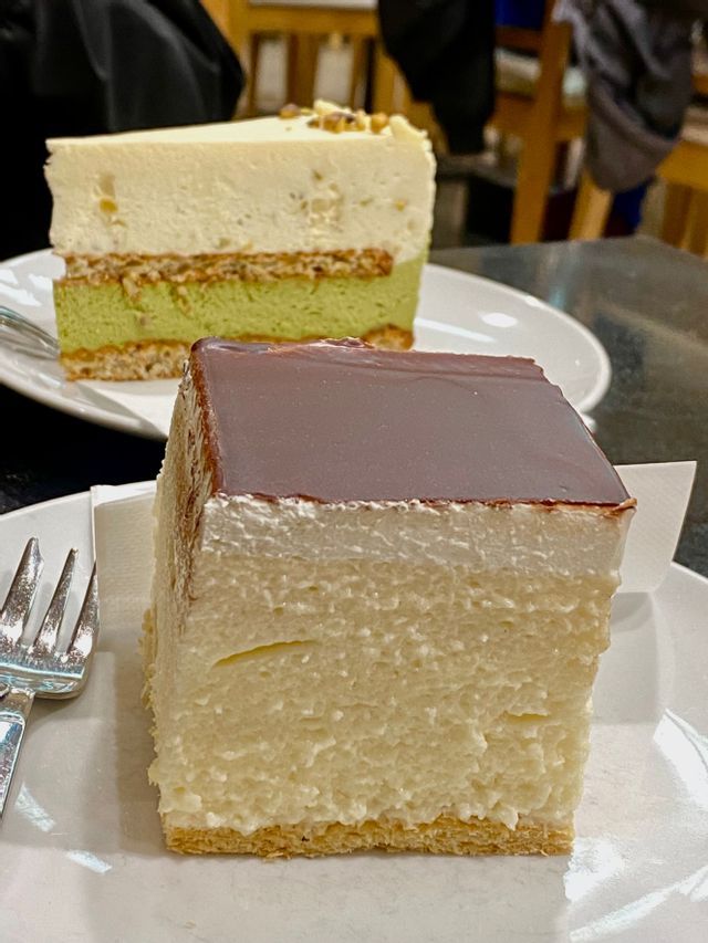 Dos rebanadas de pastel en platos blancos, con un pastel de mousse cuadrado glaseado de chocolate en primer plano y un pastel de pistacho en capas en el fondo.