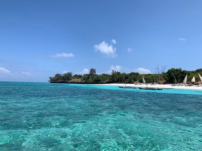Acqua turchese cristallina in primo piano con barche a vela ormeggiate su una spiaggia di sabbia bianca, una costa alberata e un cielo blu.
