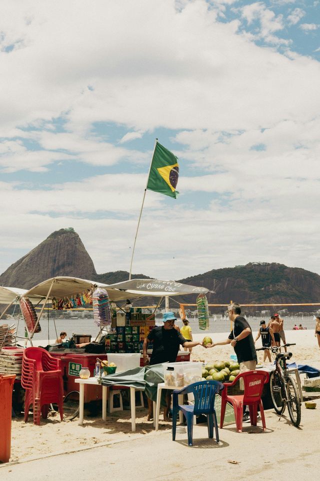 Un venditore su un carretto da spiaggia con una bandiera brasiliana serve una noce di cocco fresca a un cliente, con una montagna sullo sfondo.