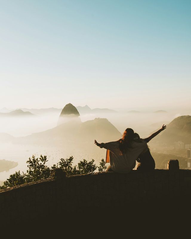 Due persone sedute su un muro con le braccia distese, che ammirano una catena montuosa nebbiosa durante un'alba dorata.