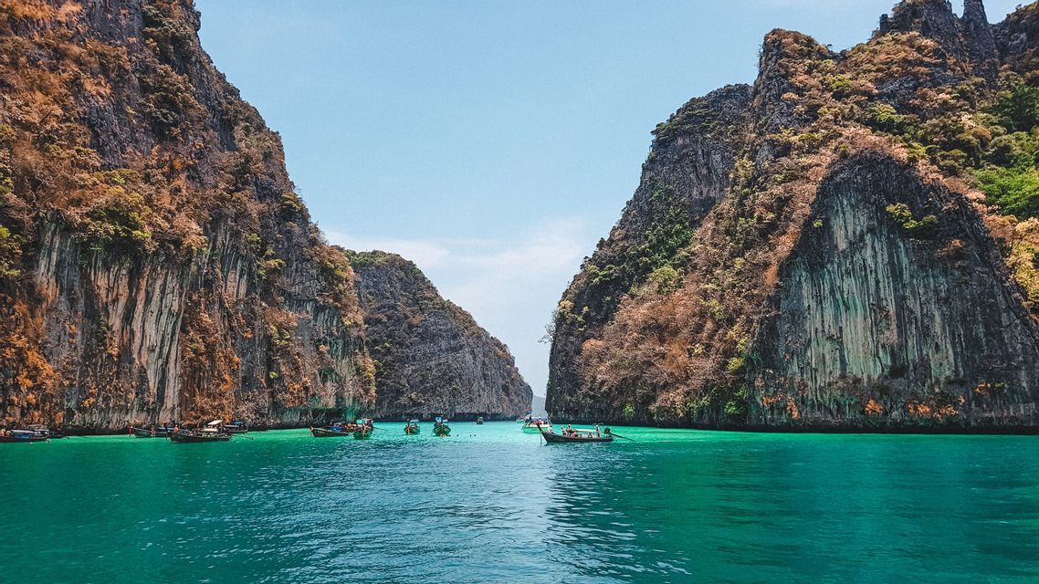 Long-tail boats float on turquoise water between two large, rocky cliffs covered in trees under a clear sky.